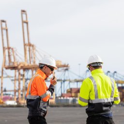 Two workers in high-visibility jackets and safety helmets communicate via radio at a busy shipping port. Cranes and cargo containers in the background indicate industrial logistics operations.